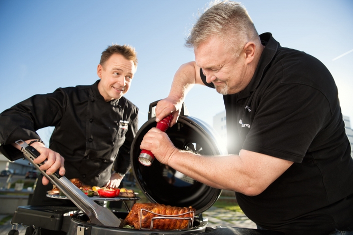 Koch und Mann stehen von einem Grill und würzen das Grillgut.